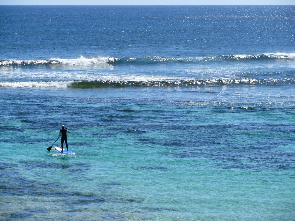 View of the back of a man in the distance, standing on a paddle board, paddling out alone into the ocean towards the waves
