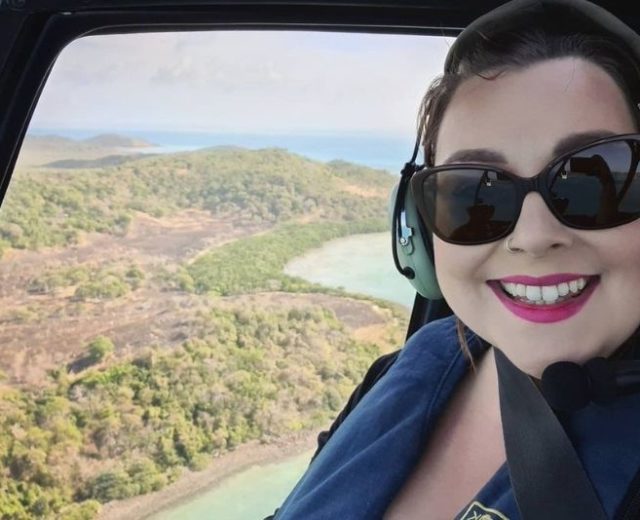 A selfie of a woman smiling while flying in a helicopter over an island and its ocean