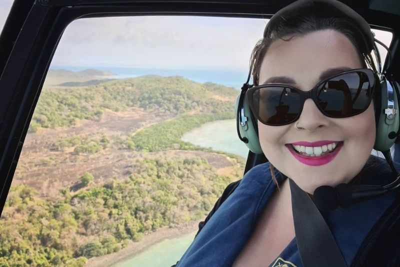 A selfie of a woman smiling while flying in a helicopter over an island and its ocean